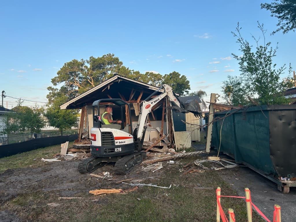 Bobcat excavator tearing down a carport structure beside a roll-off dumpster